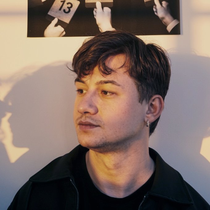 A portrait of a young man with dark wavy hair wearing a black jacket and silver hoop earring, with a black and white photograph showing hands holding the number '13' on the wall behind him.