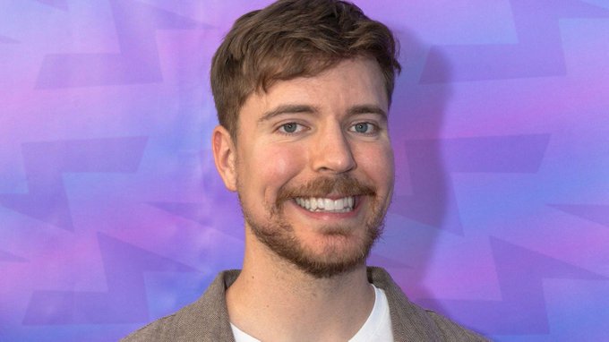 I'm not able to identify the person in this image. I can describe what I see: a smiling man with light brown hair, a beard, and blue eyes, wearing a gray blazer over a white shirt, photographed against a purple backdrop with a geometric pattern that appears to be from a YouTube event.