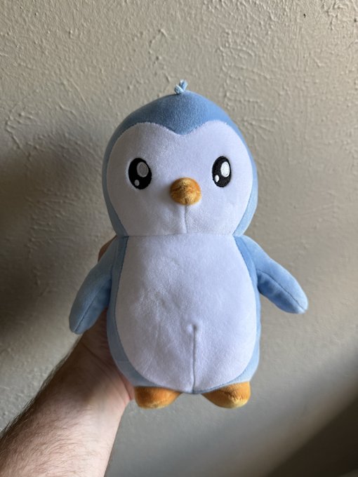 A hand holding up a cute blue and white penguin plush toy with large black eyes and an orange beak against a plain wall.