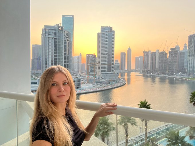 A blonde woman poses on a balcony overlooking a waterfront city skyline at sunset, with high-rise buildings, palm trees, and water in the background.