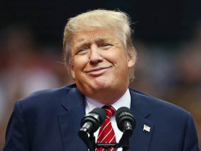 A photograph of Donald Trump smiling in a dark suit with a red striped tie, standing at a microphone against a blurred background.