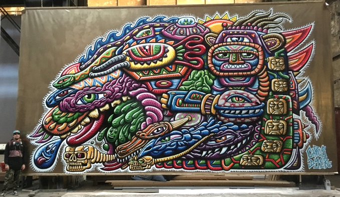 A vibrant, large-scale mural featuring intricate Aztec/Mesoamerican-inspired designs with snakes, skulls, and stylized faces in vivid colors, with an artist standing to the left for scale.