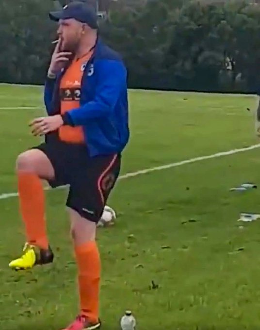 A photograph of a bearded man in a blue and orange sports kit, black cap, and yellow cleats standing on a grassy soccer field with his leg raised.