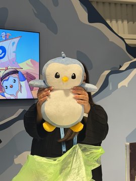 A person in a black hoodie holds a large penguin plush toy in front of their face, standing in front of a screen displaying anime artwork.