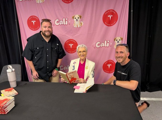 Two men posing with an elderly woman who is holding a book at a 'Cali' book signing event for 'Tesla Queens' featuring a Silicon Valley-themed backdrop.