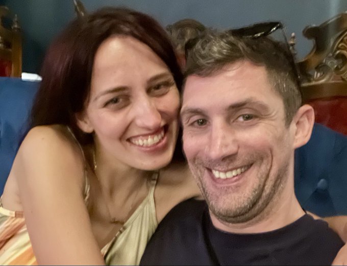 A selfie of a smiling couple—a woman with dark hair and a man with short brown hair—sitting closely together indoors.