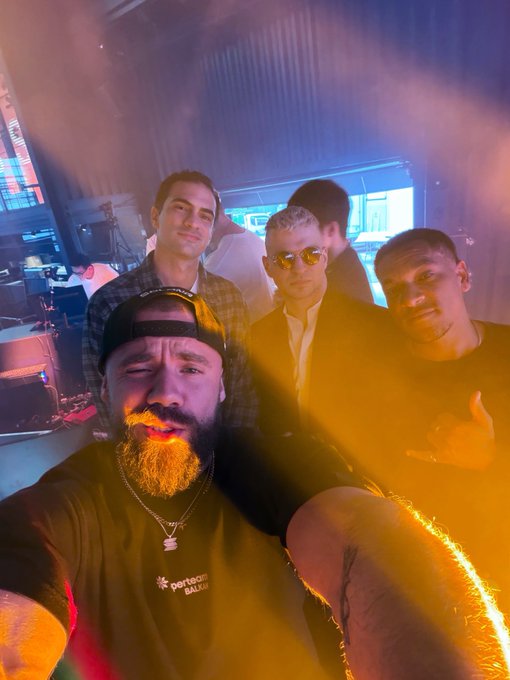 A group selfie of four young men in a dimly lit venue with stage lights and smoke; one in front has a beard dyed yellow and wears a "PortoCam BALIJA" shirt.