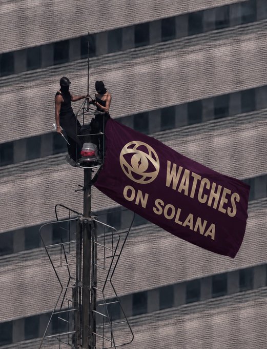 Two people dressed in black secure a large maroon banner reading "WATCHES ON SOLANA" to a flagpole atop a building, visually promoting the brand highlighted in the accompanying post.