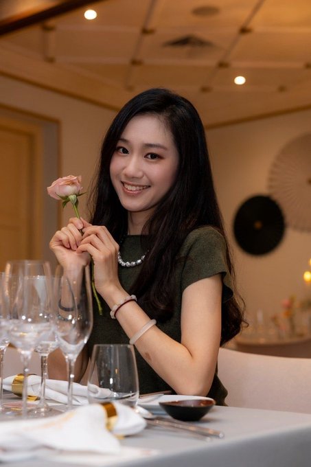 A smiling woman wearing a pearl necklace and a green top holds a pink rose while sitting at a restaurant table set with wine glasses and chopsticks.