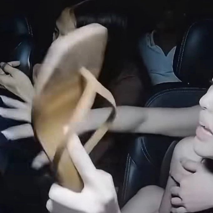 A blurry, chaotic scene taken inside a car shows a tan object appearing to strike a person's face, with another person in a white shirt visible in the back seat.