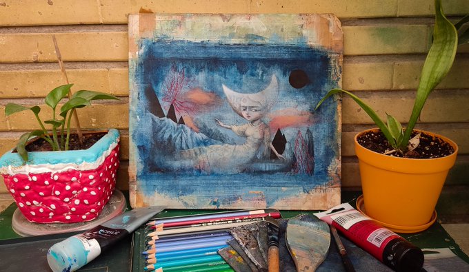 A mixed-media painting titled "Lullaby of Moon Lake" (22.19 cm) rests on an easel between potted plants, depicting a dreamy blue-toned scene of a moonlit figure floating above water with a crescent moon face, surrounded by paint tubes, brushes, and colored pencils.