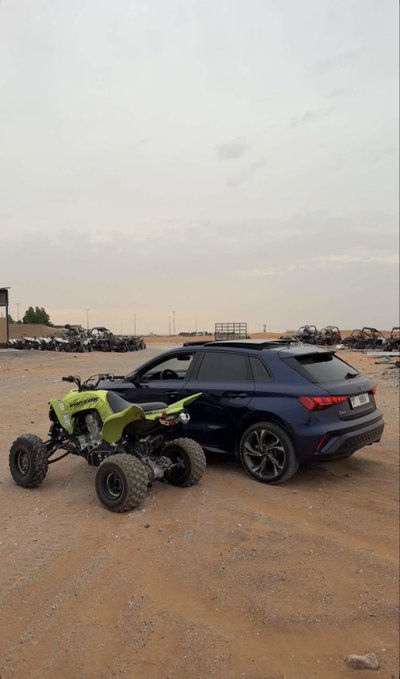A black Audi SUV parked on a dirt lot next to a green ATV, with numerous other off-road vehicles lined up in the background under a cloudy sky.