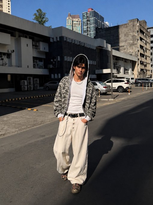 A street-style fashion photo of a young man with dark hair wearing a camouflage hooded jacket, white t-shirt, oversized white pants with chains, and brown shoes, standing on an asphalt road in an urban setting with modern high-rise buildings in the background.