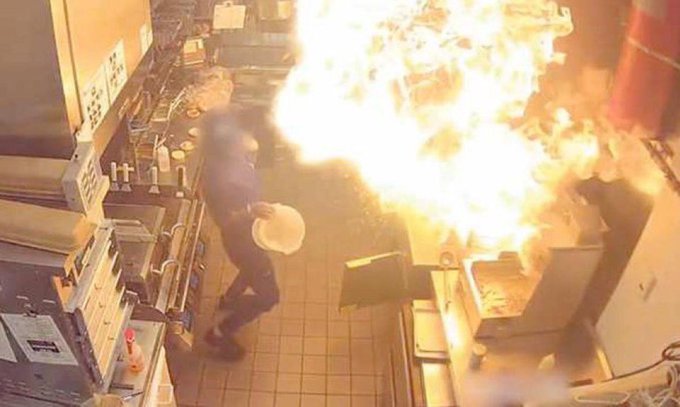 Surveillance footage captures a massive fire erupting on a stove in a commercial kitchen as a worker in a blue shirt reacts.