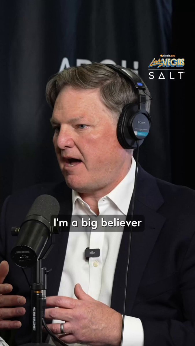 The image features a man wearing headphones, speaking into a microphone while holding a microphone stand. The background includes a banner with the text "I'm a big believer" written above him. This suggests that he might be discussing or promoting Bitcoin as a means of payment or belief in its utility and resistance to censorship.