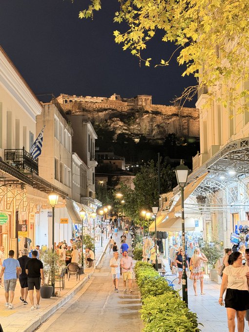 A nighttime street scene in what appears to be Athens, showing pedestrians walking along a pedestrian street lined with shops and cafes, with the illuminated Acropolis visible on the hilltop in the background.