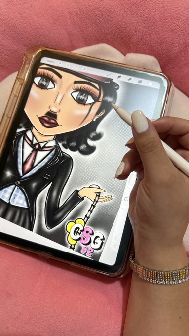 A person using a white stylus to digitally draw a stylized, anime-like female character with large eyes, dark hair, and a black suit on a tablet screen, resting on a pink surface.