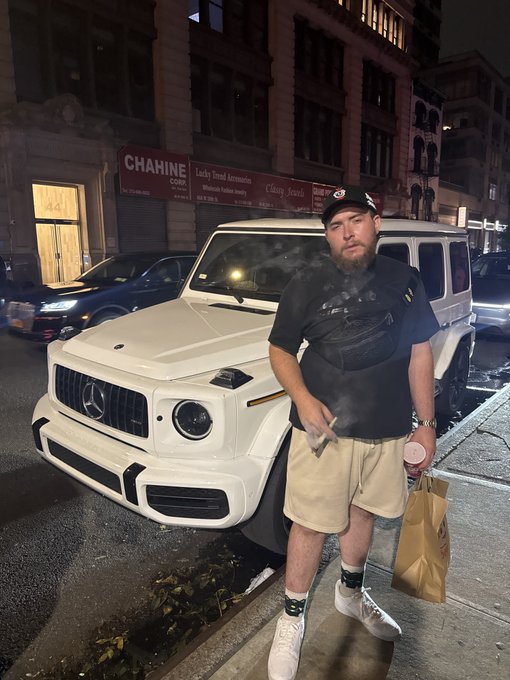 A man in a black cap, t-shirt, and khaki shorts stands on a nighttime city street in front of a white Mercedes-Benz G-Wagon, smoking with visible vapor around him.