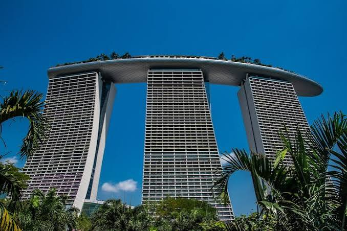 A photograph of the Marina Bay Sands hotel in Singapore, showing its three towering structures connected by a rooftop park under a clear blue sky, framed by palm fronds.
