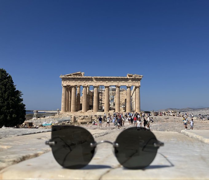 A pair of round sunglasses rests on stone in the foreground, with the ancient Parthenon temple in Athens, Greece, and tourists visible in the background.