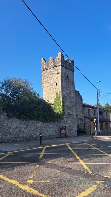 A medieval stone tower with crenellations stands against a clear blue sky, viewed from a street corner with yellow road markings and a traffic light in the foreground.