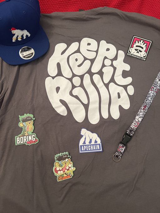 A dark gray t-shirt displays the bold bubble-letter text "KEEP IT RILLA" in cream/white, surrounded by streetwear accessories including a blue snapback hat, a graphic lanyard, and several stickers/branding elements for "BORING" and "APECHAIN" featuring ape-themed graphics—aligning perfectly with the post's hype about the "Keep It Rilla" merch drop from @ApeChainRILLAZ and @boringmerch.