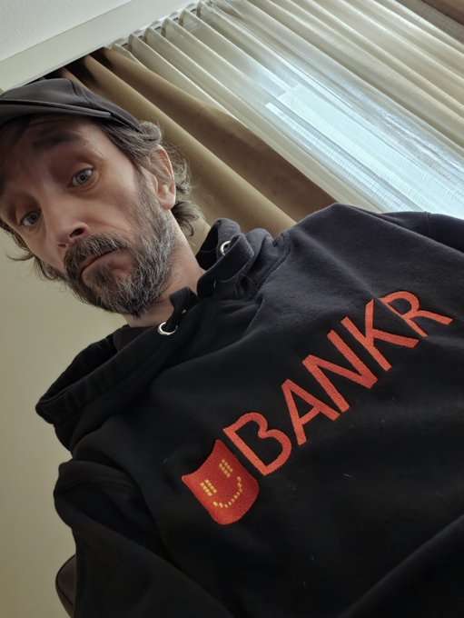 The image features a man wearing a black hoodie with the word "BNKR" printed on it, along with an orange logo. The man appears to be posing for a photo or selfie, as he has his head tilted back and is looking directly at the camera. The background of the image shows a window, suggesting that they might be indoors in a room. There are no other people visible in the picture, making it a self-portrait.
