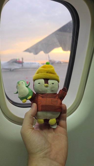 A hand holds a plush toy wearing a yellow hat and orange outfit, with a small bird plushie attached, viewed through an airplane window showing a wing and tarmac at sunset.