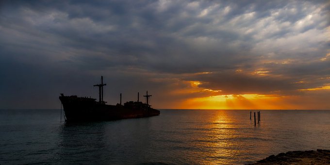 A silhouetted shipwreck sits in calm waters against a stunning orange sunset with sun rays breaking through clouds.