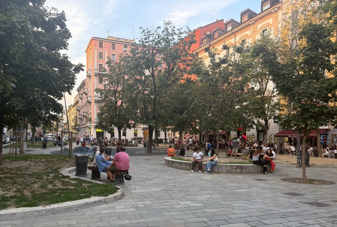 A lively European city square at dusk, with people relaxing on benches beneath trees as warm sunlight illuminates the surrounding buildings. The caption implies that none of the casually gathered individuals realize "$Umia is tradable tomorrow," suggesting a sense of unawareness amid the everyday scene.