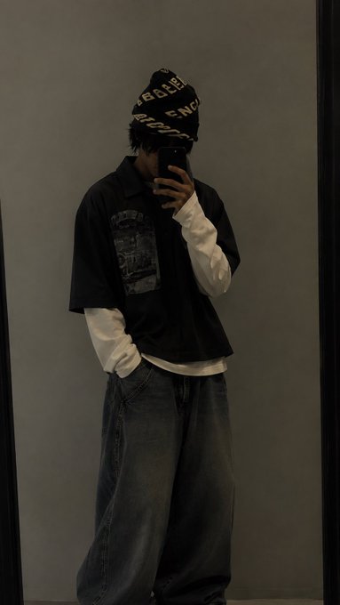 A mirror selfie of a person wearing streetwear, including a black beanie with white text, a layered black and white shirt, and loose-fitting blue jeans.