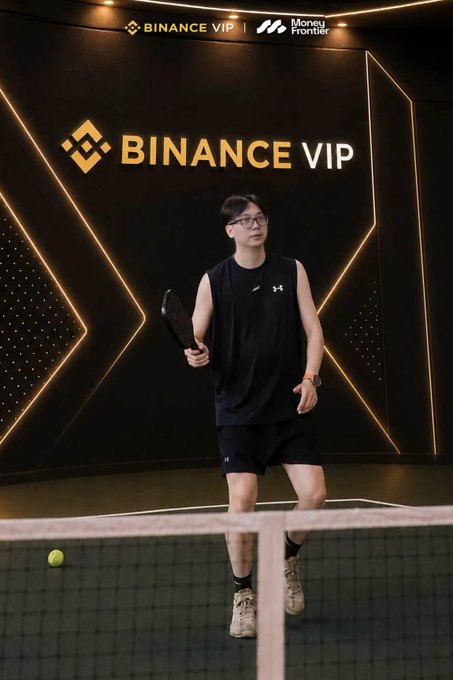 A man wearing glasses and athletic wear holding a pickleball paddle on a court, with a 'BINANCE VIP' and 'Money Frontier' branded wall in the background.