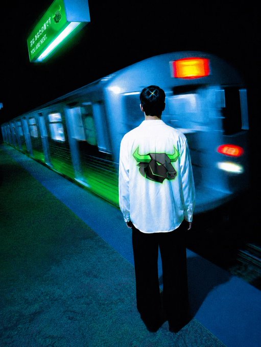 Photo of a person from behind wearing a white shirt with a green and black bull logo, standing on a train platform as a blurred train passes by under green and blue lighting.