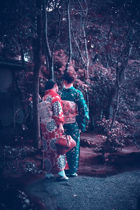 The image shows two people dressed in traditional Japanese clothing walking through a garden or park with trees around them. The person on the left has their back turned, while the other person is facing forward. Both individuals are carrying bags, possibly filled with personal belongings or souvenirs from their journey. The scene captures the essence of Japan's rich cultural heritage and its connection to nature.