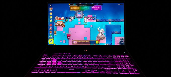 A laptop with pink RGB backlit keys displays a vibrant 2D pixel-art video game featuring colorful characters on a blue and pink background.