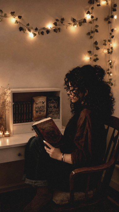 A moody, atmospheric photo of a woman with curly dark hair sitting in a chair reading a book, surrounded by warm fairy lights, ivy, candles, and a bookshelf with Harry Potter books visible.