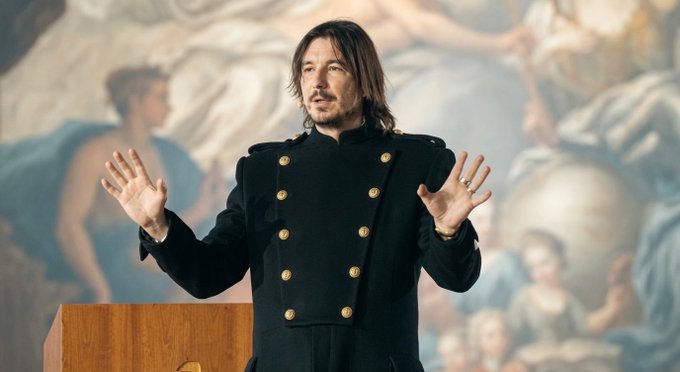 A man with shoulder-length dark hair and a goatee stands at a wooden podium, wearing a black double-breasted military-style jacket with gold buttons, gesturing with both hands raised while speaking in front of a classical painting backdrop.

The image appears to show a presenter or speaker at the Robinhood World event, likely delivering one of the international expansion announcements mentioned in the post regarding the company's expansion into the UK and Canada.