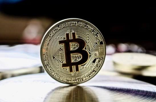 A close-up photograph of a physical gold Bitcoin coin standing upright on a reflective surface, with text around the edge including 'BITCOIN DIGITAL DECENTRALIZED PEER TO PEER'.