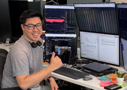 A smiling man wearing glasses and headphones around his neck gives a thumbs-up while seated at a multi-monitor trading workstation displaying financial charts, market data, and analytics dashboards. The setup reflects the post's theme of productive work during a bear market, showcasing the tools and dedication behind FalconX Global's growth and the expansion of their credit vault to $150M through partnerships with Pareto Credit and M11 Credit.