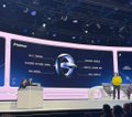 A blurry, wide shot of a brightly lit stage at an event with a large curved screen in the background and small figures of people in the foreground.