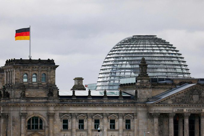 The German flag flies above the Reichstag building in Berlin, which houses the Bundestag. The visible inscription "DEM DEUTSCHEN" on the building's facade underscores Germany's national context, aligning with the announcement of Germany's proposed 25% flat tax on crypto profits.