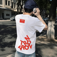 A person viewed from behind wears a white t-shirt featuring the red text 'NOW OR NEVER' on the back, along with a black cap and headphones.