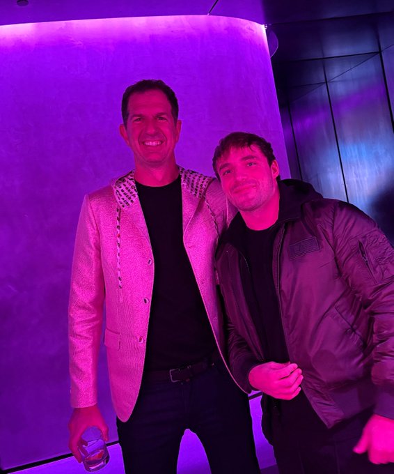 A photo of two men posing together under purple lighting, with the taller man on the left wearing a light-colored blazer with studded lapels and holding a water bottle.