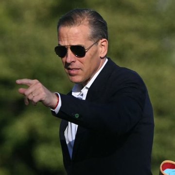 A man wearing a dark suit, white shirt, and sunglasses points off-camera with his right hand while standing outdoors against a blurred green background.