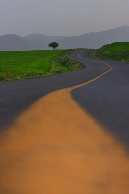 The image shows a winding road with an orange line painted on it, leading towards a mountain range in the distance. The road curves gently around the mountains, creating a sense of depth and perspective. The sky above appears cloudy, adding to the overall atmosphere of the scene. The post about rainy days is related to this image because the presence of the winding road and the orange line suggests that it might have rained recently or could rain soon. This would make the road wet and potentially more challenging for drivers due to reduced visibility and slippery conditions.