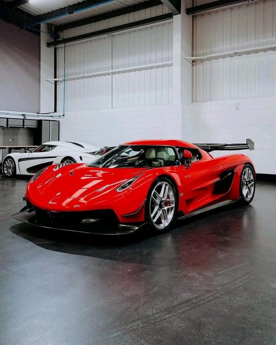 The image features a red sports car parked inside a large garage or warehouse, with another white car visible in the background. The red car appears to be an exotic sports car, while the other car seems more like a regular sedan. Both cars are parked next to each other on a concrete floor, and there is a person standing nearby.