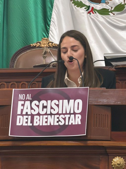 A woman speaks at a podium featuring a Mexican flag, with a prominent sign reading 'NO AL FASCISIMO DEL BIENESTAR' displayed on the lectern.