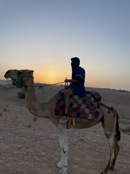 A person in a blue outfit rides a camel in a desert landscape at sunset, with a colorful patterned saddle blanket.