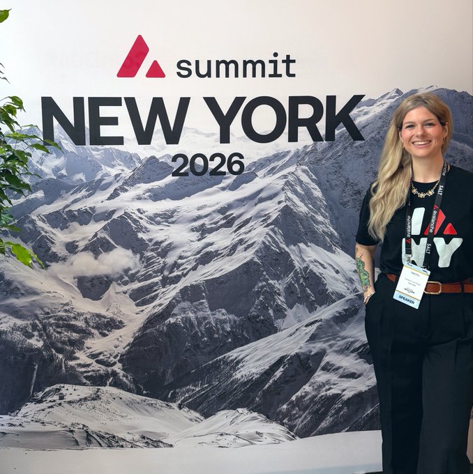 A woman with long blonde hair wearing a Summit New York 2026 t-shirt and speaker badge stands smiling in front of a backdrop featuring snowy mountains and the 'Summit NEW YORK 2026' logo.