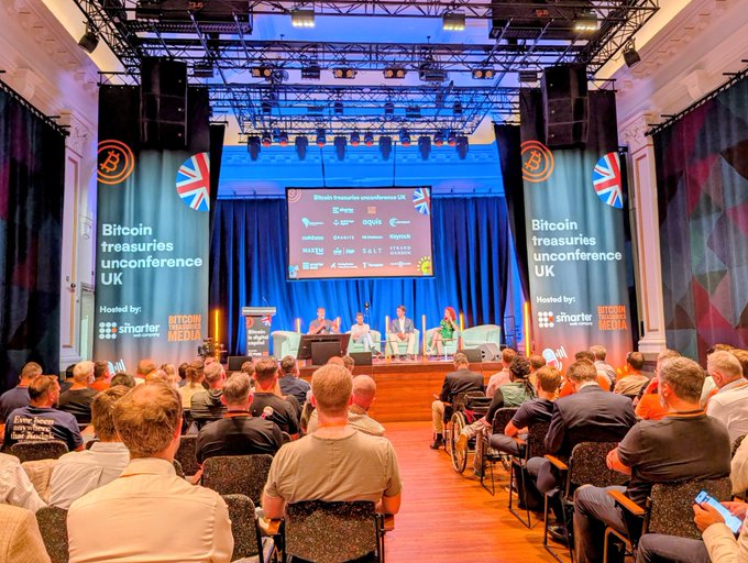 The image shows a large group of people sitting in chairs, with some standing behind a stage at an event. The stage has a blue curtain backdrop that displays information about Bitcoin and its impact on society. A few people are seated in front of the stage, while others are scattered around the room. There is also a TV screen visible in the scene.

The post related to this image highlights the Bitcoin Treasury UnConference held at Croesus Bank in London, UK. The event aims to discuss and explore the potential impact of Bitcoin on society and its implications for financial systems worldwide.
